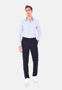 Camicia a righe blu chiaro con maniche lunghe, indossata con pantaloni blu navy e sneakers bianche. La camicia presenta un colletto con bottoni e un design aderente.