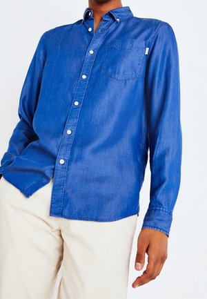 Hombre con camisa azul abotonada con bolsillo en el pecho y pantalones beige sobre un fondo blanco liso.