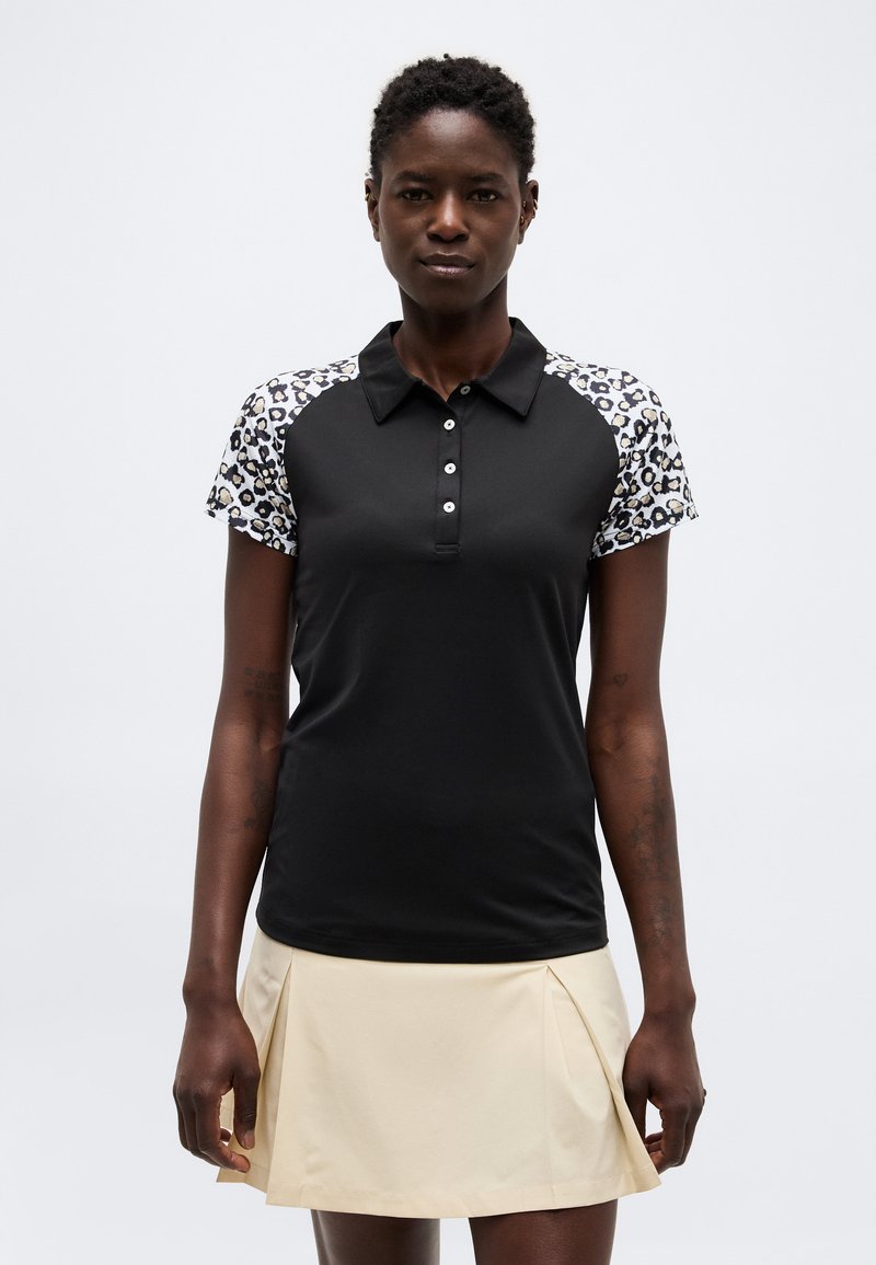 Γυναίκα που φοράει μαύρο πόλο με κοντά μανίκια με λεοπάρ μοτίβο και μπεζ πλισέ φούστα, στέκεται μπροστά σε λευκό φόντο.