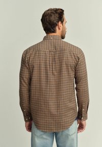 Camisa de cuadros marrón con mangas largas, cuello abotonado y dobladillo redondeado; presenta una mezcla de patrones marrones oscuros y claros.