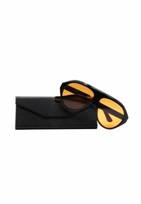 Occhiali da sole neri con montatura ottagonale e lenti arancioni, posizionati parzialmente su una custodia nera con texture.
