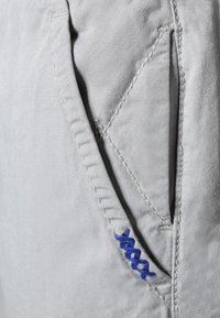 Světlé šedé tkaniny s viditelnou kapsou, která má modré švy a jemnou strukturu. Design zdůrazňuje čisté linie a praktičnost.