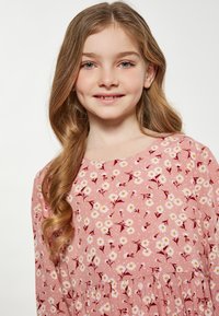 Roze bloemenjurk met witte madeliefjes en bordeauxrode accenten; lange mouwen, ronde halslijn en geplooide taille.