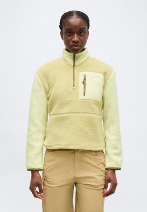 Jeune femme aux cheveux tressés portant un pull polaire jaune clair avec une poche zippée sur la poitrine et un pantalon cargo beige, debout droit.