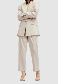 Jasny beżowy garnitur z lnu, w tym dopasowana marynarka z klapami i dwoma przednimi kieszeniami, w połączeniu z luźnymi spodniami i sandałami na obcasie.