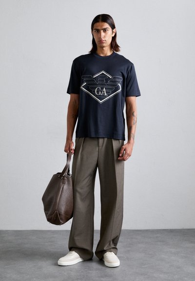 Marineblå bomulls-t-skjorte med grafisk logo i hvitt, kombinert med beige vide bukser. Holder en brun skinnveske. Hvite sneakers.