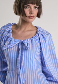 Blusa a righe blu e bianche con maniche a palloncino, scollatura arricciata e dettaglio con fiocco frontale. Realizzata in tessuto leggero.
