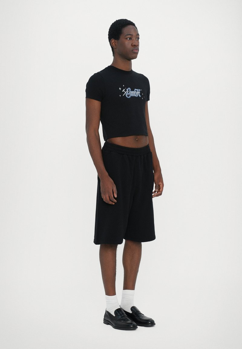 Black cropped t-shirt with graphic logo, paired with black shorts. Model wears white socks and black loafers. Simple, modern style.