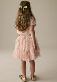Rosa Tüllkleid mit geschichteten Rüschendetails und floralen Akzenten, goldenem Taillengürtel und passendem gelbem Haarband, kombiniert mit goldenen Schuhen.