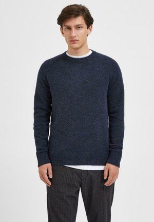 Suéter azul marino hecho de una tela suave, con un clásico cuello redondo y puños y dobladillo acanalados. Llevado sobre una camisa blanca, con pantalones oscuros.