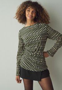 Top con maniche lunghe in verde e crema con strisce ondulate, caratterizzato da un design arricciato sul lato e un ampio scollo. Abbinato a una gonna nera.