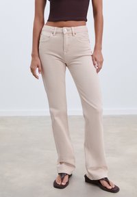 Beiga utsvängda jeans med en något texturerad yta, femficksdesign och knappstängning, i kombination med bruna flip-flops.
