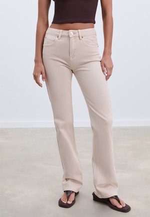 Beige Schlagjeans mit leicht strukturiertem Finish, Fünf-Taschen-Design und Knopfverschluss, kombiniert mit braunen Flip-Flops.