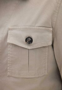 Beige stoffen overhemdzak met een klep die wordt vastgehouden door een enkele donkerviergaatjesknop, gedetailleerd met zichtbare stiksels rond de randen.