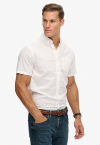 Wit button-up overhemd met korte mouwen van linnen, met een borstzak en gele knopen, gecombineerd met blauwe jeans en een gevlochten bruine riem.