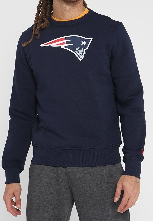 Hombre vestido con sudadera azul marino con el logo de los New England Patriots en el pecho y pantalones de chándal grises, de pie con las manos relajadas a los lados.