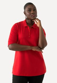 Camisa polo roja con cuello, mangas cortas y tapeta de botones. Tejido suave, con aberturas en los laterales y un ajuste ligeramente holgado.