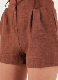 Shorts en lin marron avec une finition texturée, dotés de plis sur le devant et d'une fermeture à bouton. Le design est ajusté et arrive au milieu de la cuisse.