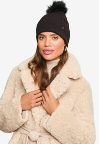 Gorro de punto negro con un pompón en la parte superior, con textura acanalada y un pequeño acento dorado. Llevado con un abrigo beige de pelaje.