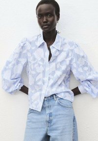 Blouse bleu clair avec un motif floral, design boutonné, manches bouffantes, associée à un jean bleu clair sur fond blanc.