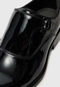 Mustan patent-nahkakengän pyöristetty varvas, sileä pinta, sivuhihna soljella ja hienovaraiset tikkausmuotoilut reunoilla.