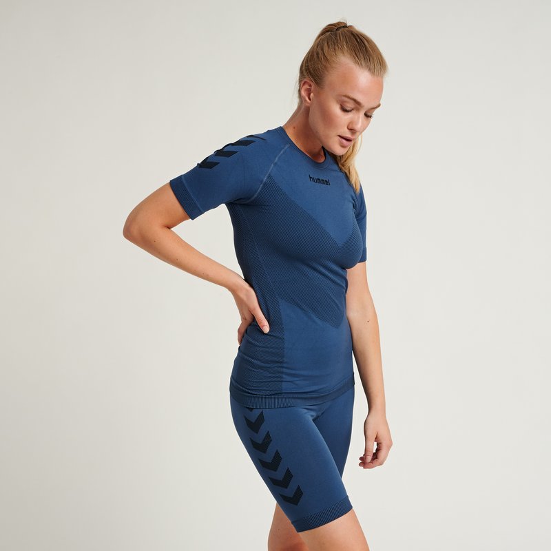 Donna con maglietta sportiva blu e pantaloncini con strisce a chevron nere, in piedi con una mano sulla parte bassa della schiena e guarda verso il basso.