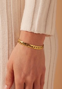 Gouden kettingarmband met in elkaar grijpende schakels, met een gepolijste afwerking. Draagt om een pols en complementair aan een gebreide trui in een lichte kleur.
