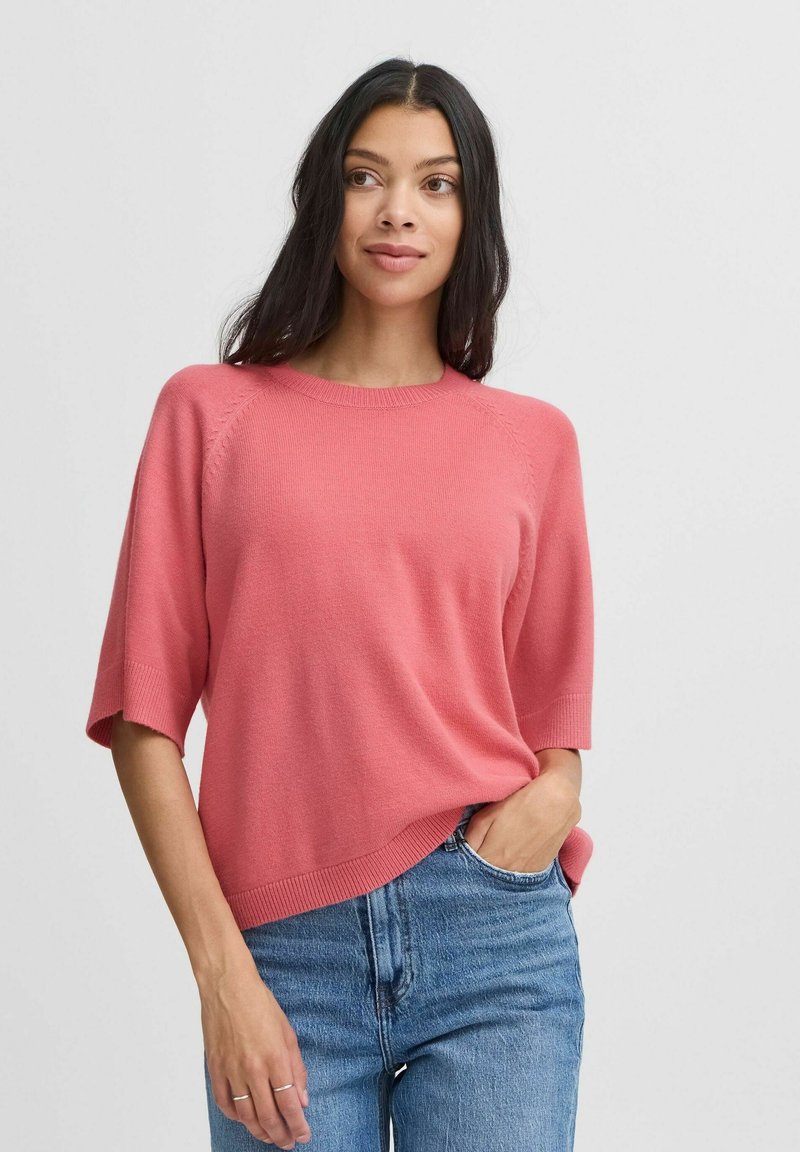 Roze gebreide trui met een ronde hals en korte mouwen, voorzien van geribde manchetten en zoom, gecombineerd met blauwe spijkerbroek.