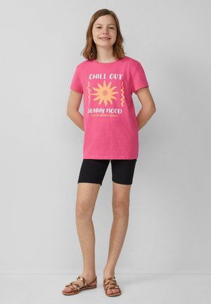 Rosa Baumwoll-T-Shirt mit dem Aufdruck "Chill Out Sunny Mood", ausgestattet mit einem gelben Sonnendesign. Kombiniert mit schwarzen Shorts und braunen Sandalen.