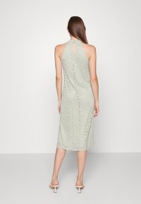 Vestido verde de renda com decote halter, detalhe em forma de olho na parte de trás, comprimento até a metade da panturrilha e corte justo. Usado com sandálias de salto em tom claro.