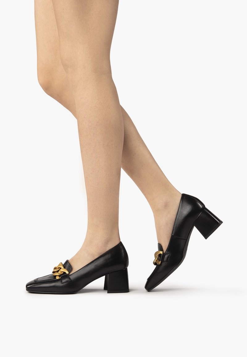 Jambes portant des mocassins à talons carrés en cuir noir avec détail de chaîne dorée, un pied à plat, l'autre relevé sur la pointe.