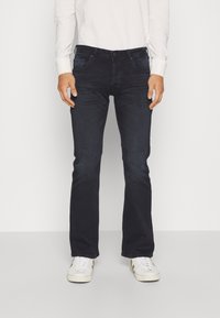 Jeans oscuros de denim para hombre, con un diseño de pierna recta, cinco bolsillos y cierre de botón. Combinados con zapatillas blancas.