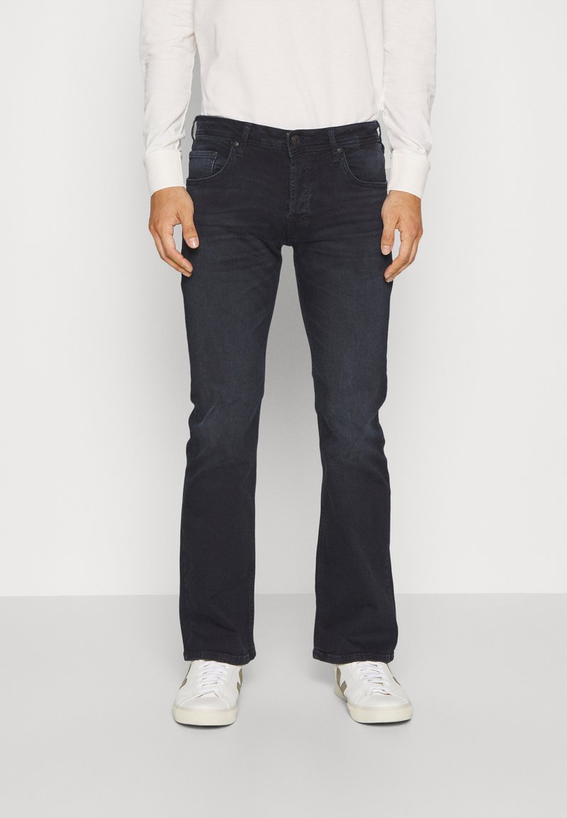 Jean bleu foncé pour homme en denim, avec une coupe droite, cinq poches et une fermeture à bouton. Associé à des baskets blanches.