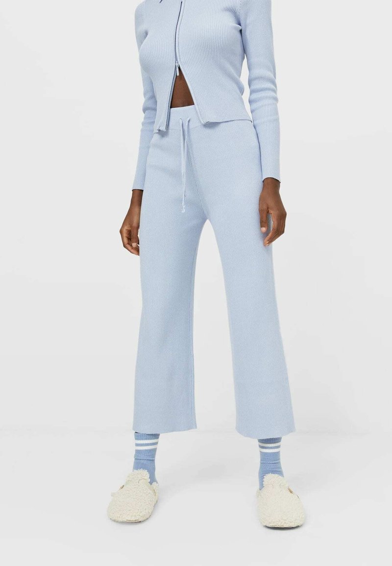 Personne portant un pantalon en maille côtelée bleu clair, un haut zippé assorti, des chaussettes bleues rayées et des chaussons pelucheux crème, debout sur un sol blanc.