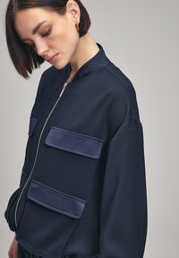 Giacca bomber della marina realizzata in tessuto liscio, con zip frontale, colletto rialzato e due tasche a pattina rettangolari sul petto.