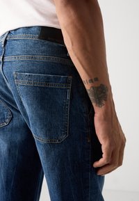 Blugi din denim albastru închis cu o croială dreaptă, având cusături contrastante, două buzunare pe spate și un eticheta din piele neagră pe talie.