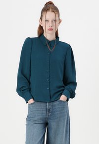 Teal button-up shirt met lange poffende mouwen en een kraag, gemaakt van textuurstof. Gecombineerd met losse blauwe denim jeans.