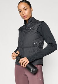 Črna puhovka brez rokavov z zadrgo spredaj, karo vzorcem in odsevnimi poudarki. Črna majica z dolgimi rokavi in bordo legice. Drži črno steklenico za vodo.