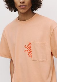 Tricou din bumbac piersici cu buzunar față și broderie portocalie în formă de corali pe partea stângă. Mâneci scurte și guler rotund.
