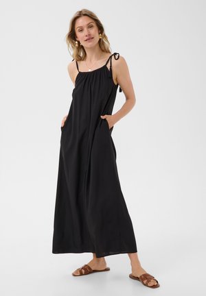 Femme portant une longue robe noire sans manches avec des bretelles à nouer et des sandales plates marron, debout les mains dans les poches de la robe sur un fond uni.