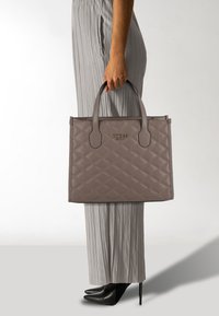 Grijze gewatteerde handtas met dubbele handvatten en ritsafsluiting, voorzien van een logo-detail aan de voorkant. Vergezeld door grijze geplooide broek en zwarte hakken.
