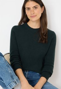 Stickad mörkgrön tröja med ribbad textur och rund halsringning. Bärs tillsammans med ljusblå denimjeans, vilket skapar en avslappnad look.