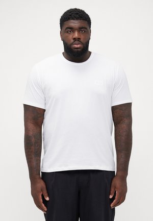 Weißes T-Shirt mit kurzen Ärmeln aus Baumwolle, runder Ausschnitt und einem kleinen, dezenten Markenzeichen auf der Brust. Texturierte Haut und sichtbare Tattoos.