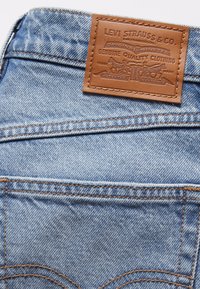Light blue denim jeans featuring a brown leather patch with embossed logo and design details. Double-stitching along the waistband.
