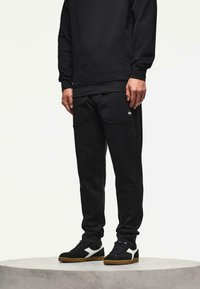 Man in zwarte joggerbroeken en zwarte sneakers met witte accenten, staande op een ronde, lichtgrijze podium tegen een effen achtergrond.