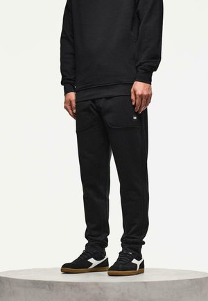 Man in zwarte joggerbroeken en zwarte sneakers met witte accenten, staande op een ronde, lichtgrijze podium tegen een effen achtergrond.