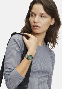 Grüne Uhr mit rundem Zifferblatt und silbernem Edelstahlband. Getragen an einem Handgelenk, kombiniert mit einem enganliegenden, grauen, gerippten Langarmshirt.
