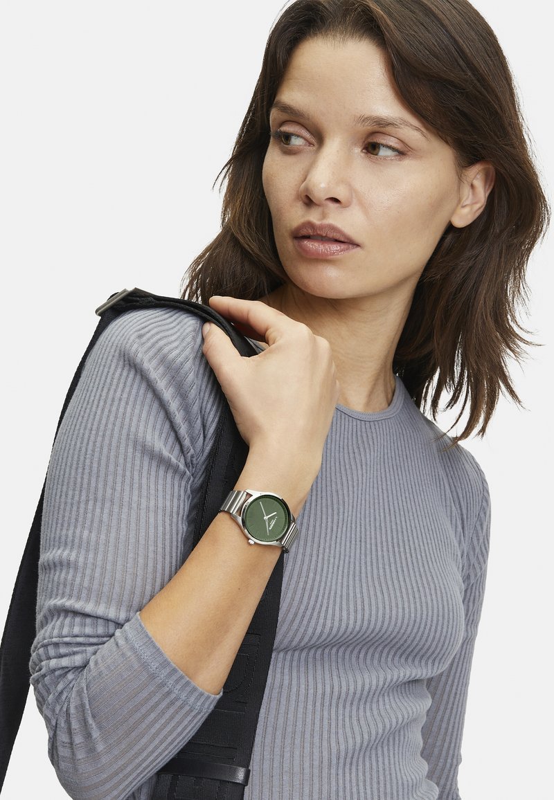 Grüne Uhr mit rundem Zifferblatt und silbernem Edelstahlband. Getragen an einem Handgelenk, kombiniert mit einem enganliegenden, grauen, gerippten Langarmshirt.