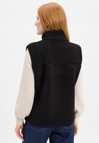 Zwarte fleecedeken met hoge kraag, mouwloos ontwerp, gestructureerde afwerking en contrasterende beige lange mouwen eronder. Effen spijkerbroek.