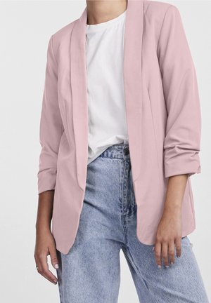 Femme portant un blazer rose pâle par-dessus une chemise blanche et un jean taille haute bleu clair, avec les manches remontées de façon décontractée.